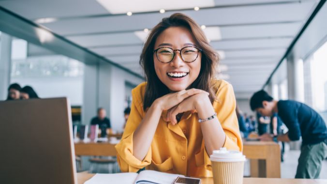 Smiling woman sitting at a laptop with an open book and beverage.